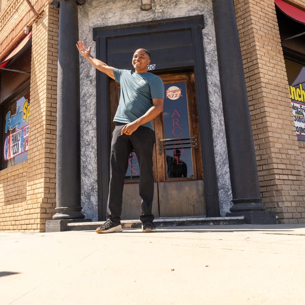 A man in a blue shirt waves in front of Corsaro's Family Pizza restaurant entrance.