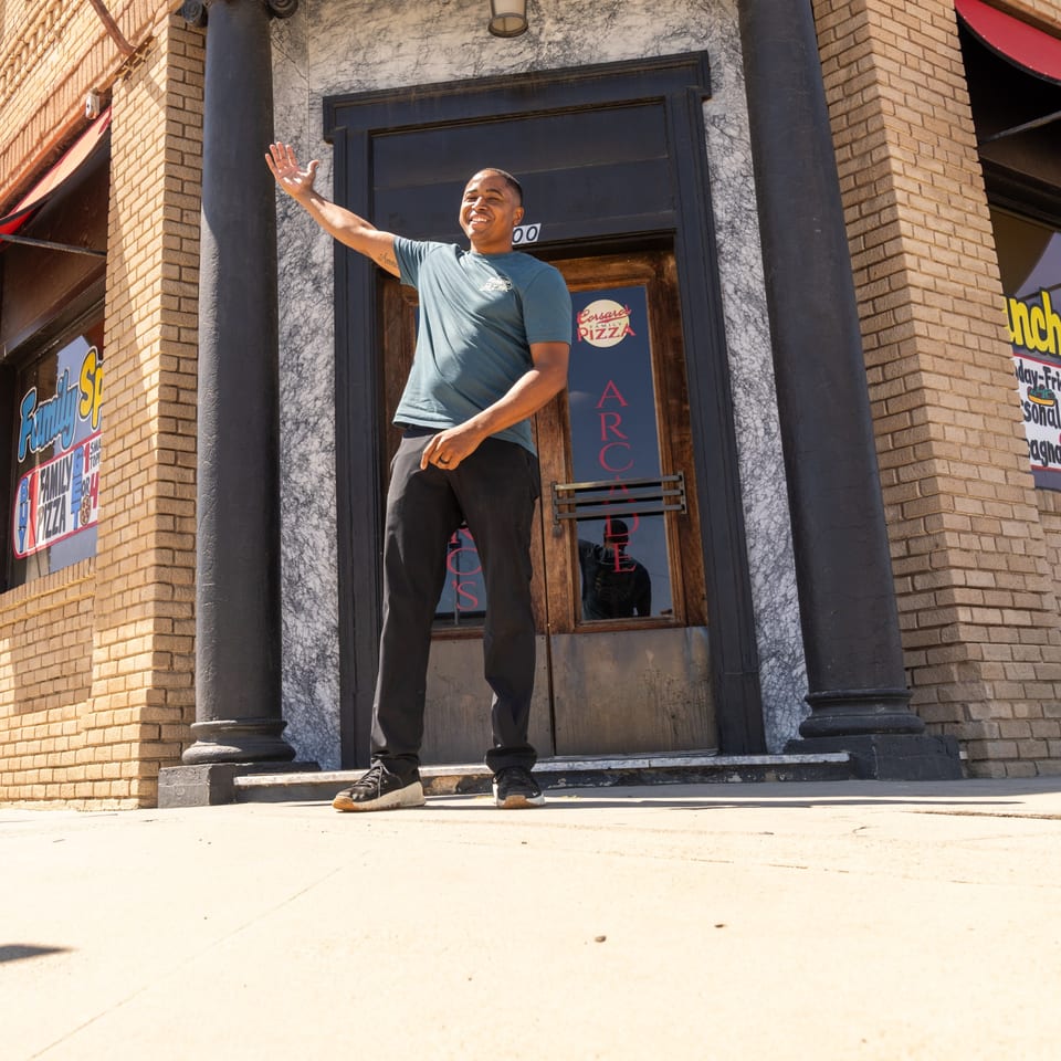 A man in a blue shirt waves in front of Corsaro's Family Pizza restaurant entrance.