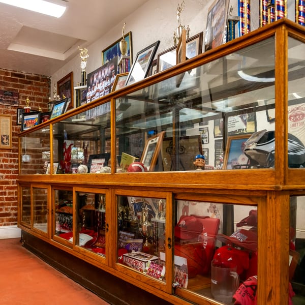 A wooden display case at Corsaro's Family Pizza filled with sports trophies, memorabilia, and photos.