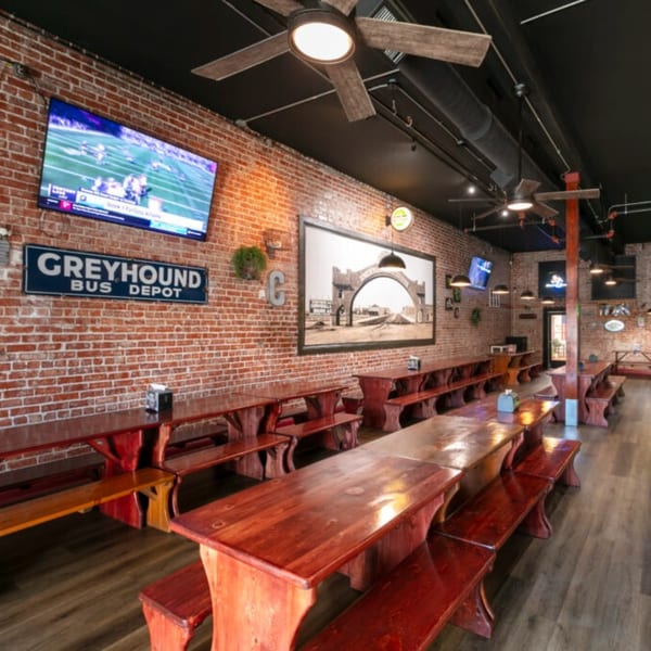Interior of Corsaro's Family Pizza with long wooden tables, benches, brick walls, and TVs showing sports.